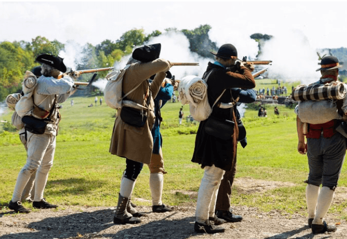 Five people in historical costumes fire muskets during a reenactment on the grassy field at Fort Ticonderoga, smoke billowing from their guns. They carry rolled blankets and backpacks, while others watch in the distance.